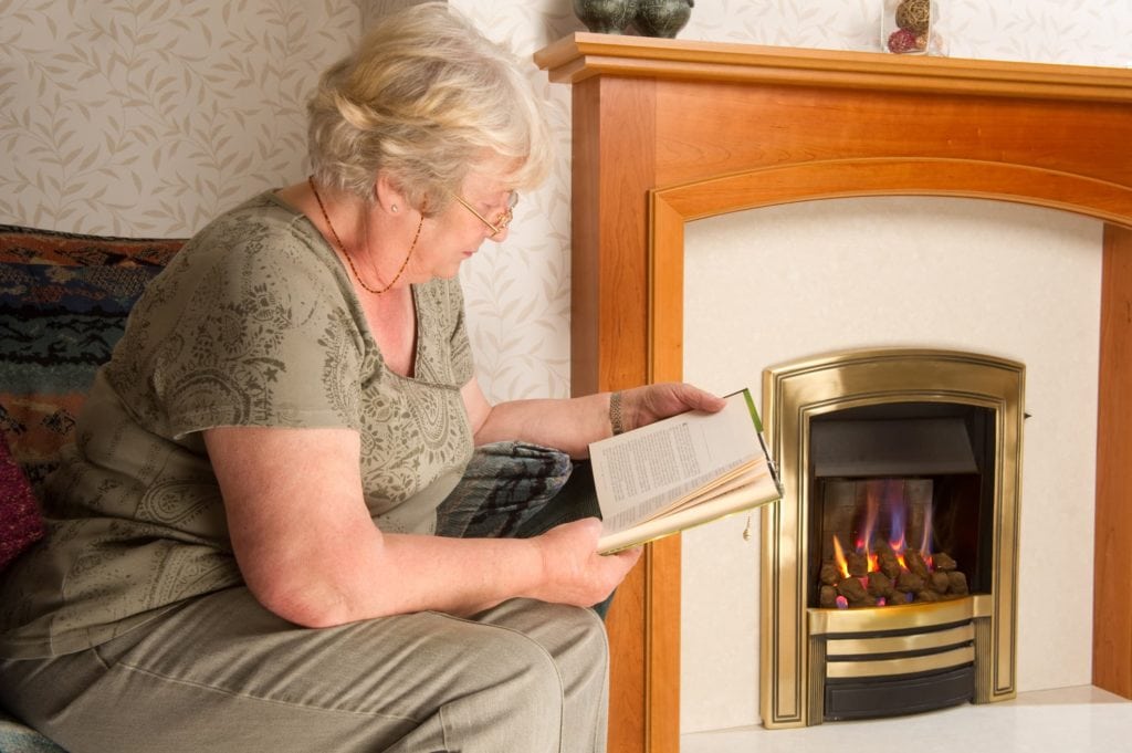 Senior woman reads a novel next to a fireplace in her home.