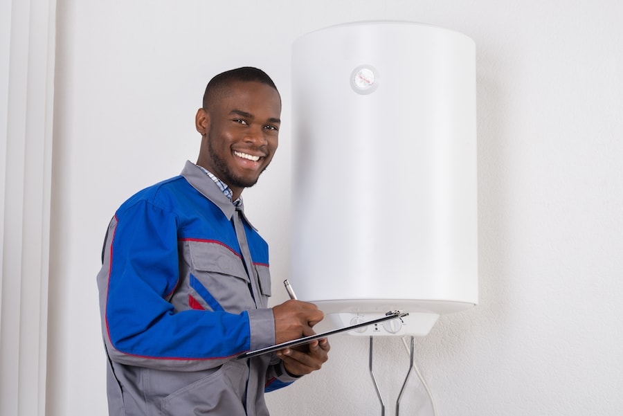 Young African Male Plumber Holding Clipboard Looking At Electric Boiler.